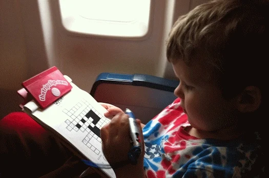 Kid on airplane doing a crossword puzzle on a red, kid-sized clipboard with attached pen