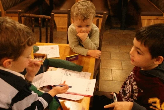Three kids at a restaurant holding pens using a kid-sized clipboard and many activity pages